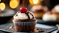 Chocolate cupcake topped with vanilla frosting, chocolate drizzle and maraschino cherry against blurred golden bokeh lights. Fresh homemade dessert on dark plate.