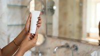 Female hand with medium skin tone holding white cosmetic tube in modern bathroom setting, ideal for skincare and beauty product marketing.