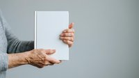 Senior adult male with aged hands holding blank white book or notebook against neutral gray background for publishing and education concepts.