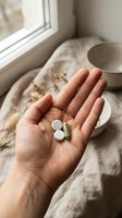 Hand holding white tablets and green capsule with ceramic cup and natural linen fabric on windowsill, representing wellness, supplements, and healthcare routine for medical and lifestyle content.