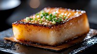 Glazed pork belly slice with caramelized surface, garnished with green onions and sesame seeds on dark slate plate for gourmet restaurant menu presentation.