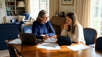 Senior businesswoman mentoring young female colleague at home office desk with laptop and documents in cozy living room setting.