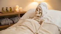 Skeleton wearing spa headwrap lying in bed with candles and towels, representing relaxation, wellness, and humorous spa treatment concepts for health and beauty marketing.