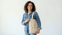 Woman with curly dark hair wearing denim shirt holding organic cotton tote bag against white background for retail and lifestyle marketing.