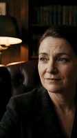 Mature Caucasian businesswoman in dark blazer sitting in cozy office interior with warm lighting and bookshelves in background for corporate portraits.