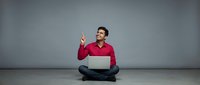 Young Hispanic man in red sweater sitting cross-legged with laptop pointing up having eureka moment against gray studio background for business concepts.