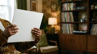 Person holding blank white paper sheet in cozy home library interior with wooden bookshelf and warm lighting, suitable for mockup and advertising projects.