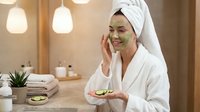 Young Asian woman applying green avocado face mask in modern spa bathroom wearing white bathrobe and towel for skincare wellness routine.