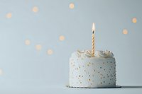 Small white birthday cake with gold sprinkles and single lit candle on light blue background with bokeh lights, minimal celebration composition.
