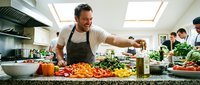 Cheerful Caucasian male chef in apron seasoning colorful fresh vegetables in a busy professional kitchen with colleagues cooking in background.