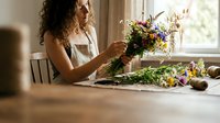 Young woman with curly hair arranging colorful wildflowers into bouquet at wooden table by window. Natural light florist workspace with fresh blooms for creative projects.