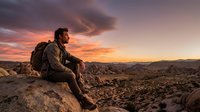 Adult male hiker with backpack sitting on rocky outcrop overlooking vast desert landscape at sunset. Warm golden and orange sky with dramatic clouds. Suitable for travel, adventure, and outdoor lifestyle content.