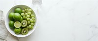 Green kiwi slices, limes and grapes arranged in white bowl on marble surface with glass and linen napkin, overhead flat lay composition for food styling and culinary content.