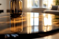 Amber glass vase on glossy black surface creating dramatic reflection with warm sunlight streaming through windows, modern minimalist home interior setting.