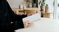 Woman in black sweater reading white letter or document at wooden table in cozy home interior with plants and ceramic decor in background.