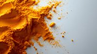 Ground turmeric powder scattered on white surface, bright yellow-orange organic spice with rich texture and small granules creating natural pattern and shadows.
