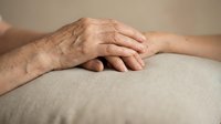 Elderly hands gently clasped together showing wrinkles and age spots on neutral fabric surface conveying comfort, care, and human connection in senior years.