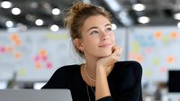Young professional businesswoman thinking with hand on chin in modern office workspace with colorful sticky notes on wall behind her.
