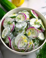 Fresh cucumber salad with sliced red onions and dill in creamy yogurt dressing, served in white ceramic bowl on marble surface. Bright summer appetizer.