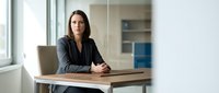 Professional businesswoman sitting at modern office desk with laptop, confident female executive in dark blazer at workplace with natural lighting.