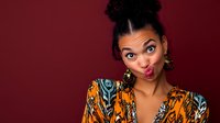 Young African American woman making funny face, wearing colorful ethnic pattern dress against burgundy background. Natural curly hair styled in bun. Playful expression.