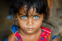 Young girl with striking blue eyes and dark hair wearing floral dress, looking directly at camera with intense gaze. Natural light portrait captures innocence and emotion.
