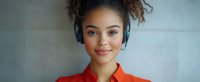 Young mixed race woman with curly hair wearing blue headphones and orange shirt smiling at camera against light background, expressing joy and confidence.