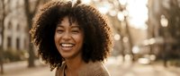 Happy young African American woman with curly hair smiling outdoors in warm sunlight, expressing joy and positivity in urban park setting.