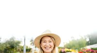 Mature woman with blonde hair wearing straw hat smiling outdoors in garden setting with flowers and greenery, suitable for lifestyle and gardening content.
