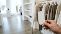 Person holding blank white tag in clothing boutique with neutral tone garments on rack, retail shopping and fashion merchandising concept.