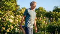 Active senior Caucasian man walking outdoors among blooming flowers in summer, enjoying healthy lifestyle and nature in warm sunlight.