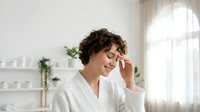 Young woman with short curly brown hair and fair complexion applying skincare product to face in bright bathroom with shelves and plants.