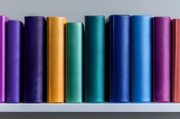 Row of colorful hardcover books arranged on white shelf, spines showing vibrant spectrum from purple through yellow to blue and pink, creating minimalist library display.