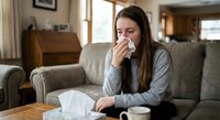 Young Caucasian woman with long brown hair sitting on couch blowing nose with tissue, feeling unwell at home during cold or flu season.