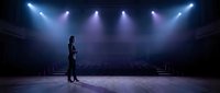 Young woman performer standing alone on empty theater stage under dramatic spotlights with audience seating visible in background, creating atmospheric scene.