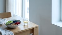 Wooden table with fresh fruit bowl containing kiwi and pomegranate seeds, glass of water, and wooden chair by bright window with white curtains in minimalist interior.