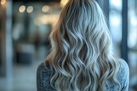 Platinum blonde wavy hair styled with soft curls on woman wearing gray sweater, back view with blurred bokeh background. Professional hairstyle and coloring.