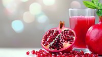 Fresh pomegranate cut in half showing juicy red seeds next to glass of bright pomegranate juice and whole fruit on light bokeh background with mint garnish.