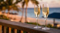 Two champagne flutes filled with sparkling wine on wooden railing overlooking tropical beach at sunset, romantic celebration vacation concept.