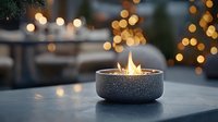 Cozy outdoor fire bowl with dancing flames against blurred holiday lights background creates warm ambiance on concrete patio table for evening entertaining.