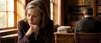 Mature woman with gray hair and Caucasian ethnicity wearing dark blazer, sitting thoughtfully with hand near chin in warm-lit office or study room with wooden furniture and windows, suitable for business, professional, corporate, and contemplative themes.