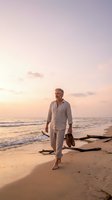 Middle-aged man with gray hair and beard walking barefoot on sandy beach at sunset, wearing light linen shirt, holding shoes, ocean waves and driftwood visible, golden hour lighting.