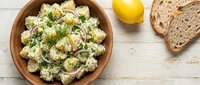 Creamy potato salad with fresh herbs and lemon served in wooden bowl on white rustic table, overhead view for food styling and recipe content.