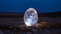 Glowing translucent egg-shaped crystal orb illuminated against desert landscape at twilight with deep blue sky creating mystical atmospheric scene.