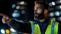 Middle Eastern construction supervisor in safety vest pointing at night, illuminated by bokeh lights against dark background. Professional leadership at worksite.