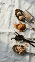 Aromatic baking spices arranged on linen fabric with natural shadows. Nutmeg, cinnamon, vanilla beans and brown sugar in wooden spoons for culinary recipes.