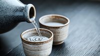 Traditional Japanese sake being poured from ceramic tokkuri bottle into handcrafted ochoko cups on wooden table creating ripples in clear rice wine.