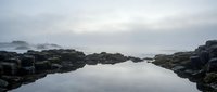 Serene tidal pool surrounded by dark rocks reflects overcast sky, creating mirror-like water surface. Coastal landscape for nature and travel content.