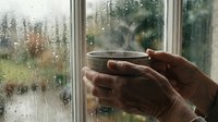 Hands holding warm coffee cup by rainy window with water droplets creating cozy indoor atmosphere during stormy weather for relaxation and comfort.