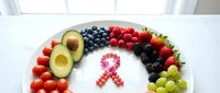 Fresh colorful fruits and vegetables arranged in rainbow pattern around pink ribbon symbol on white plate for healthy eating awareness campaign.
